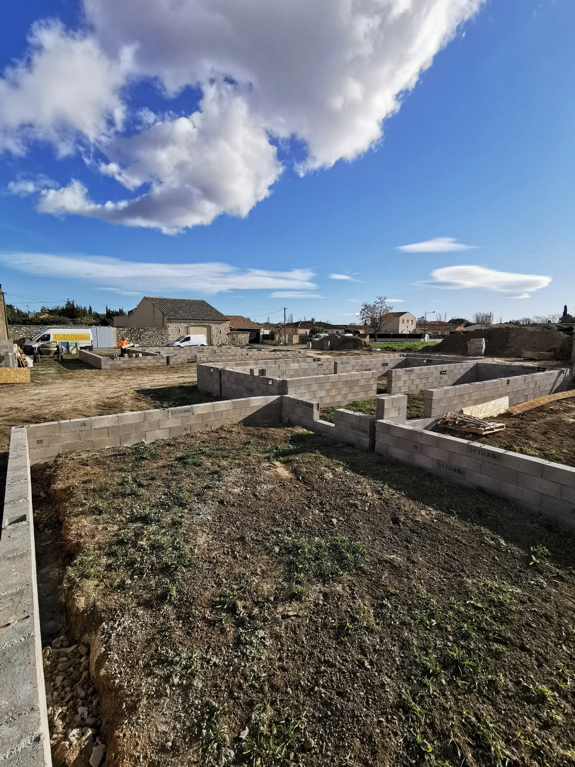 Chantier de construction avec fondations en parpaings et vide sanitaire sous un grand ciel bleu nuageux.