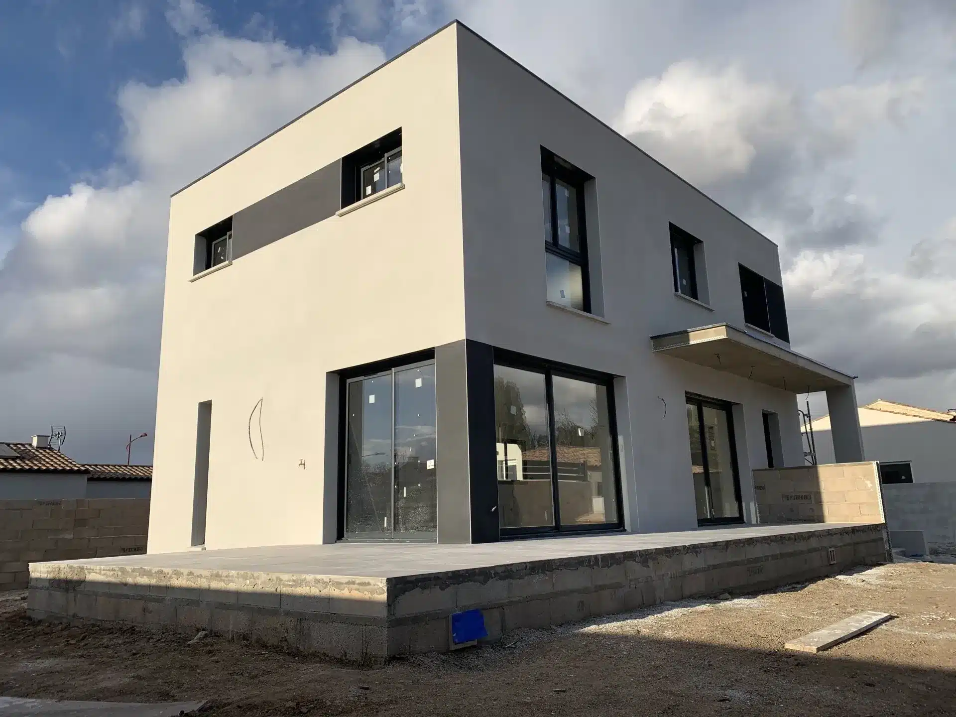 Maison cubique moderne bicolore en fin de chantier avec larges baies vitrées sur terrasse bétonnée.