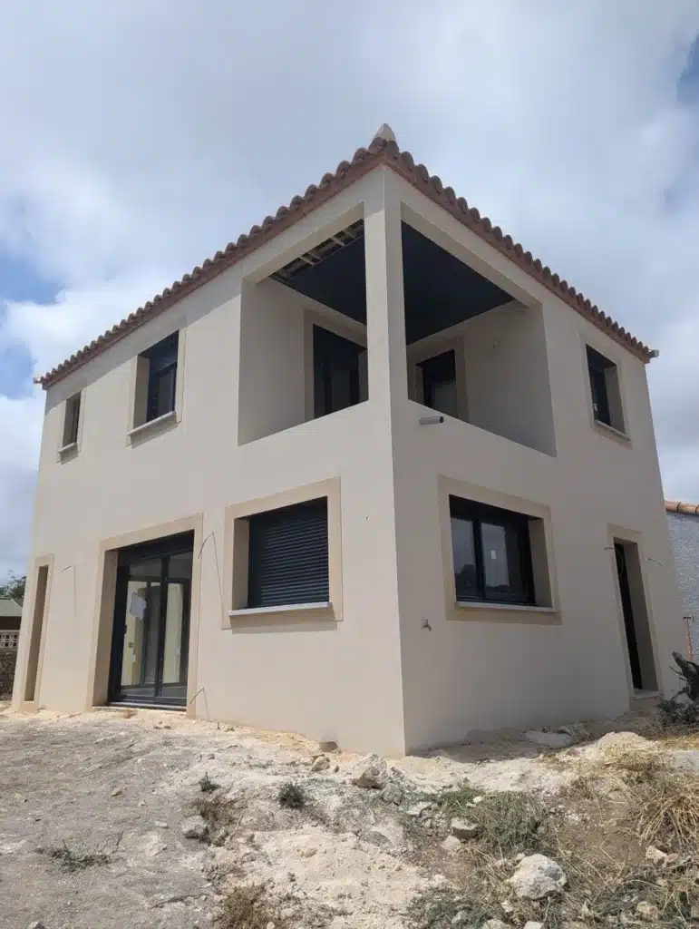 Maison neuve beige à deux étages avec menuiseries noires, terrasse couverte et toit en tuiles sous un ciel nuageux.