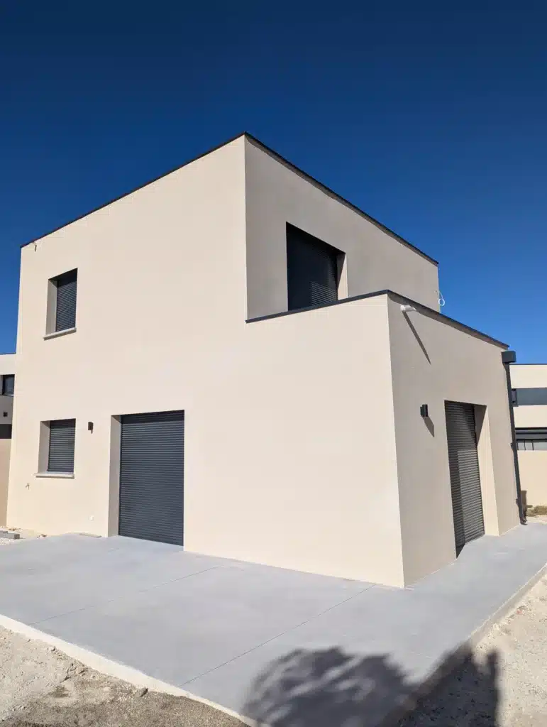 Maison cubique moderne à façade beige et volets roulants gris anthracite sur terrasse en béton sous un ciel bleu.